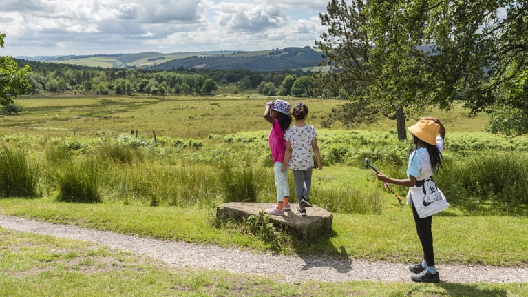 Two children wearing sun hats standing on a small rock next to a path looking out over the countryside with another child standing on a path next to them.